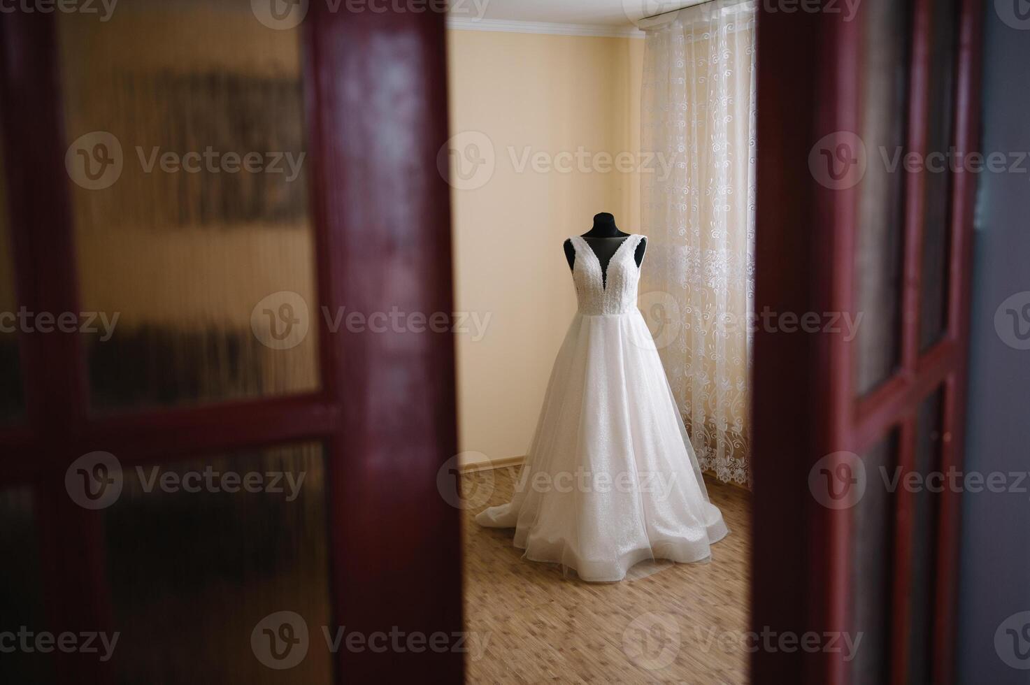 Beautiful bride's white wedding dress hangs near the bed in a hotel room with flowers at the bottom. Bridal morning before ceremony photo