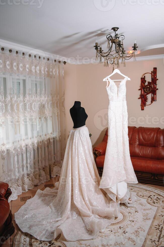 Beautiful bride's white wedding dress hangs near the bed in a hotel room with flowers at the bottom. Bridal morning before ceremony photo