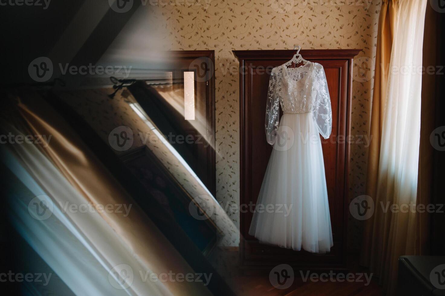 Beautiful bride's white wedding dress hangs near the bed in a hotel room with flowers at the bottom. Bridal morning before ceremony photo