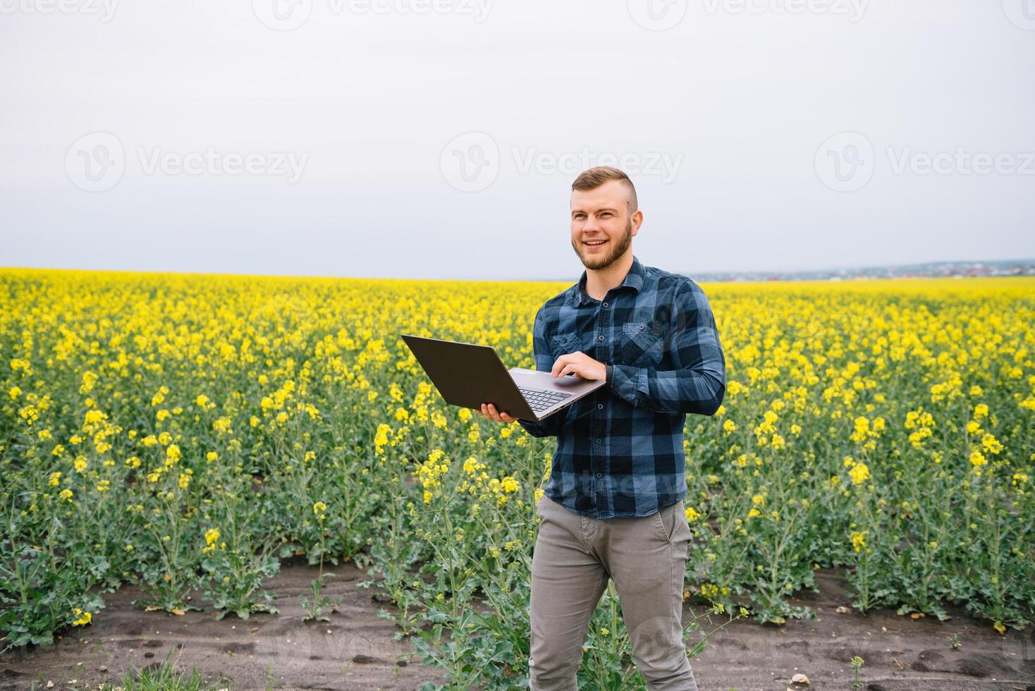 joven agrónomo sostiene ordenador portátil en violación campo. agronegocios concepto. agrícola ingeniero en pie en un violación campo con un ordenador portátil en verano. foto