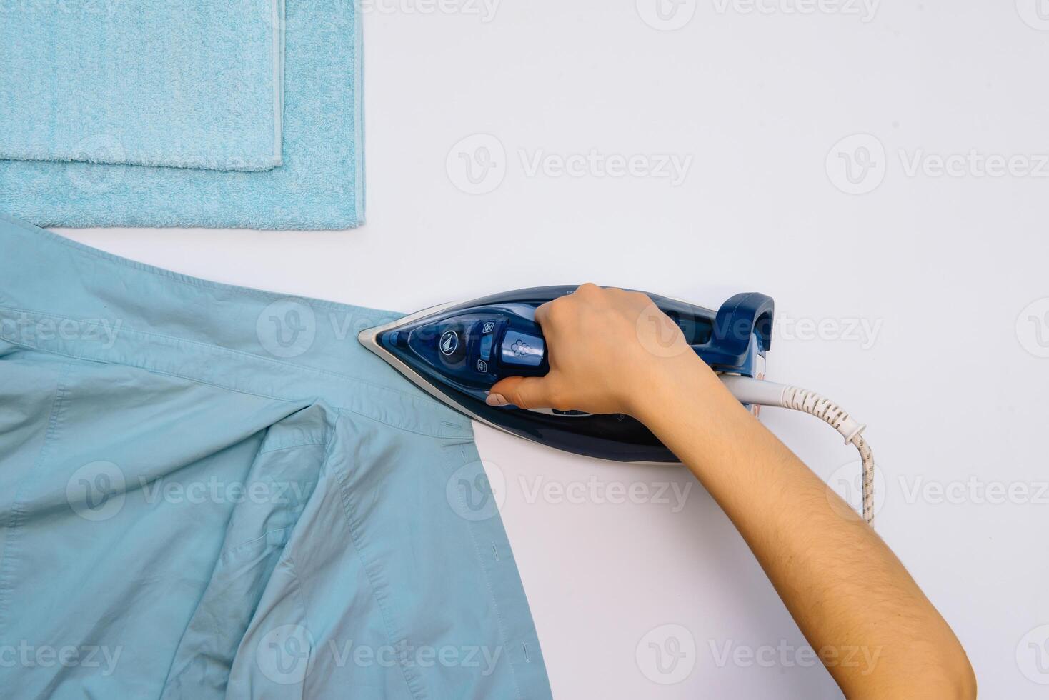 Female hand ironing clothes top view isolated on white background. Young woman with iron ironing man's shirt seen from above during housework. Blue iron on white table photo