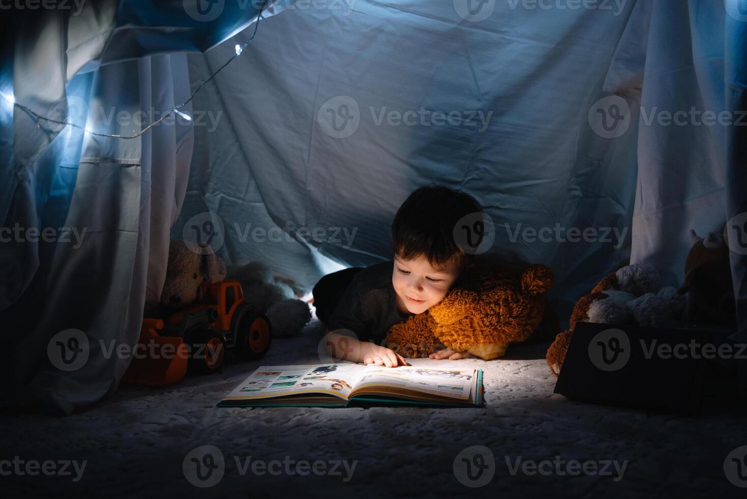 niño chico leyendo con libro y Linterna y osito de peluche oso en carpa. antes de yendo a cama. foto