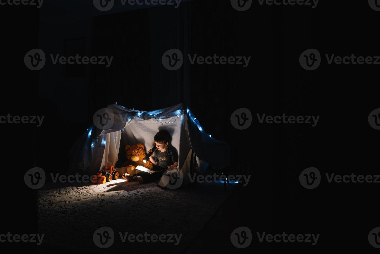 child boy reading with book and flashlight and teddy bear in tent. before going to bed. photo