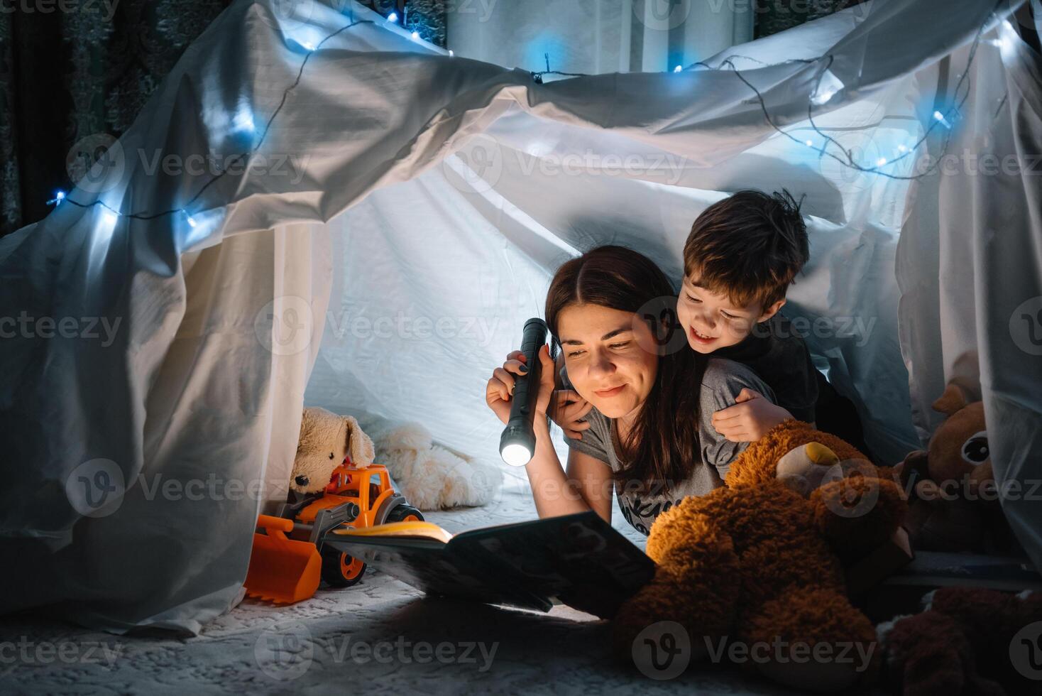 happy family mother and child son reading a book with a flashlight in a tent at home. family concept. photo