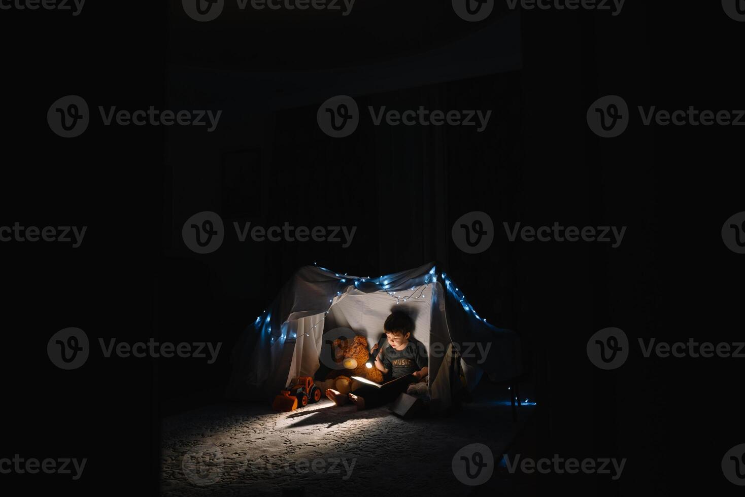 child boy reading with book and flashlight and teddy bear in tent. before going to bed. photo