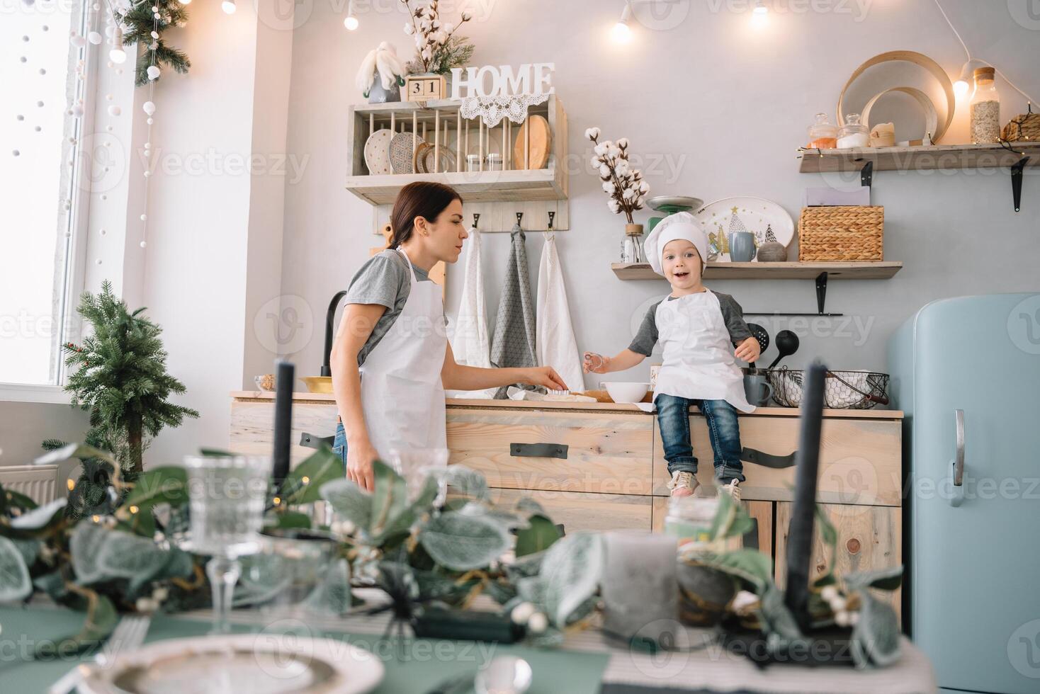 joven contento mamá y su bebé cocinar galletas a hogar en el cocina. Navidad hecho en casa pan de jengibre. linda chico con madre en blanco uniforme y sombrero cocido chocolate galletas. foto