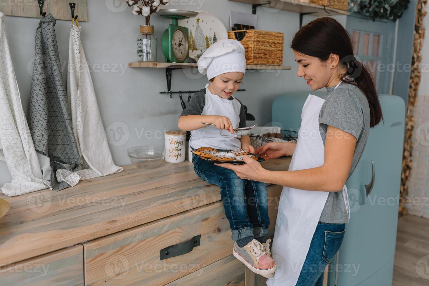 joven contento mamá y su bebé cocinar galletas a hogar en el cocina. Navidad hecho en casa pan de jengibre. linda chico con madre en blanco uniforme y sombrero cocido chocolate galletas foto