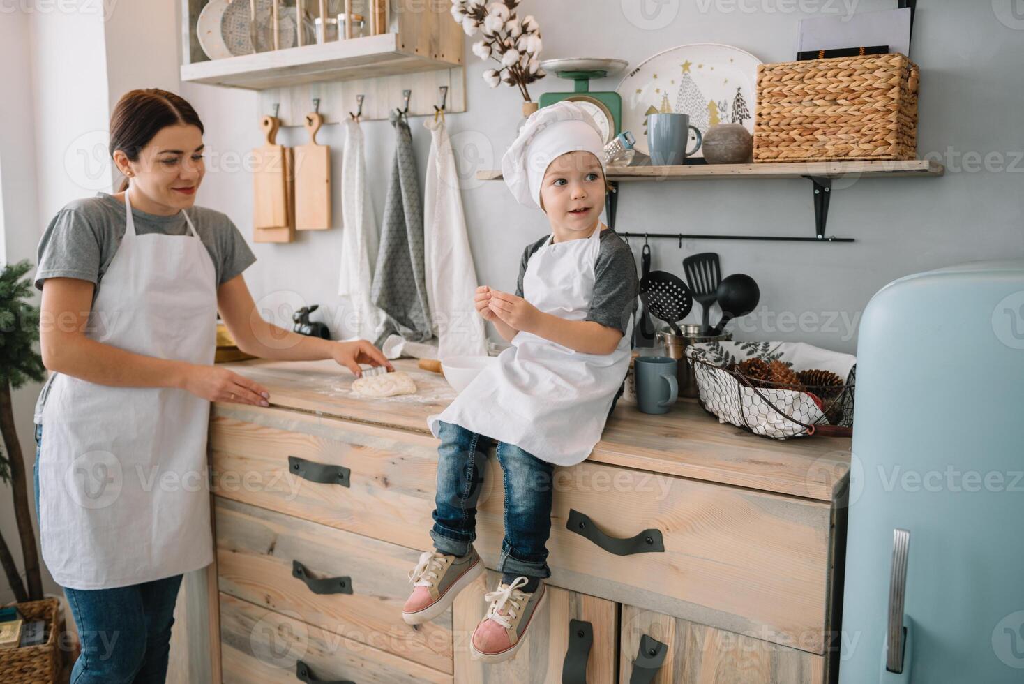 joven contento mamá y su bebé cocinar galletas a hogar en el cocina. Navidad hecho en casa pan de jengibre. linda chico con madre en blanco uniforme y sombrero cocido chocolate galletas. foto