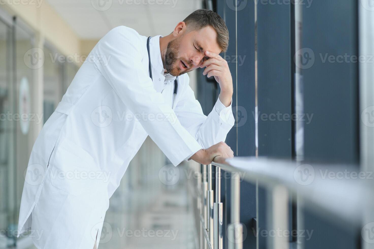 Sad doctor leaning against the wall in hospital corridor. photo