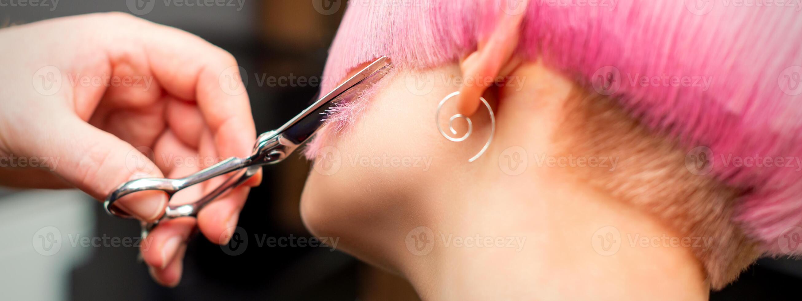 Cutting female hair. Hand of a hairdresser cutting short pink hair of young white woman at the hair salon. photo