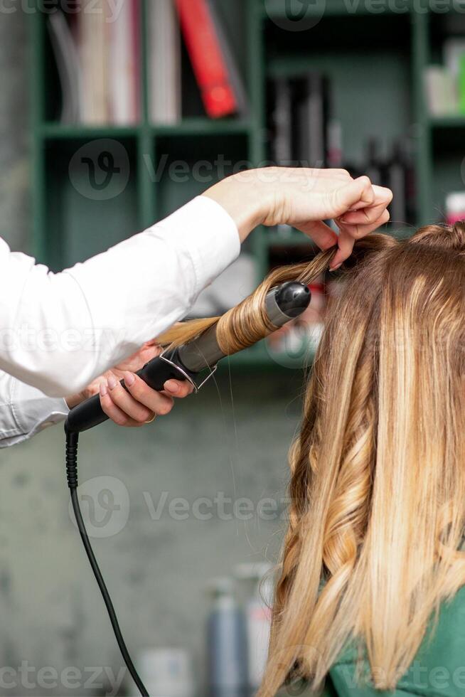 Creating curls with curling irons. Hairdresser makes a hairstyle for a young woman with long red hair in a beauty salon. photo