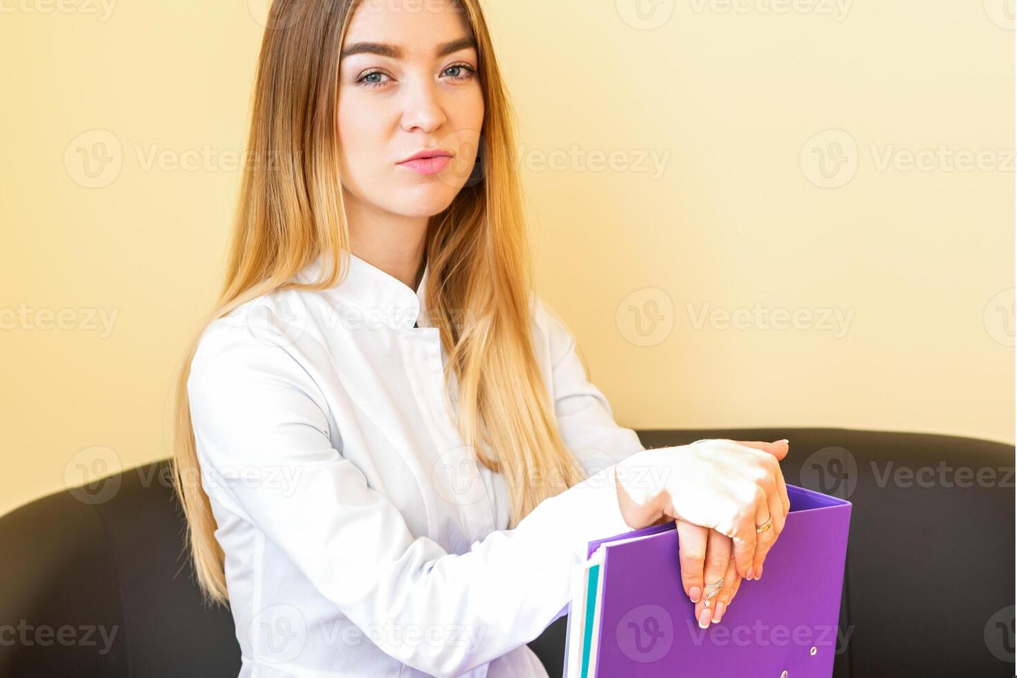 The young healthcare professional, dentist, doctor, nurse, the assistant in a white coat with folders sitting on a black sofa and looking at the camera. photo