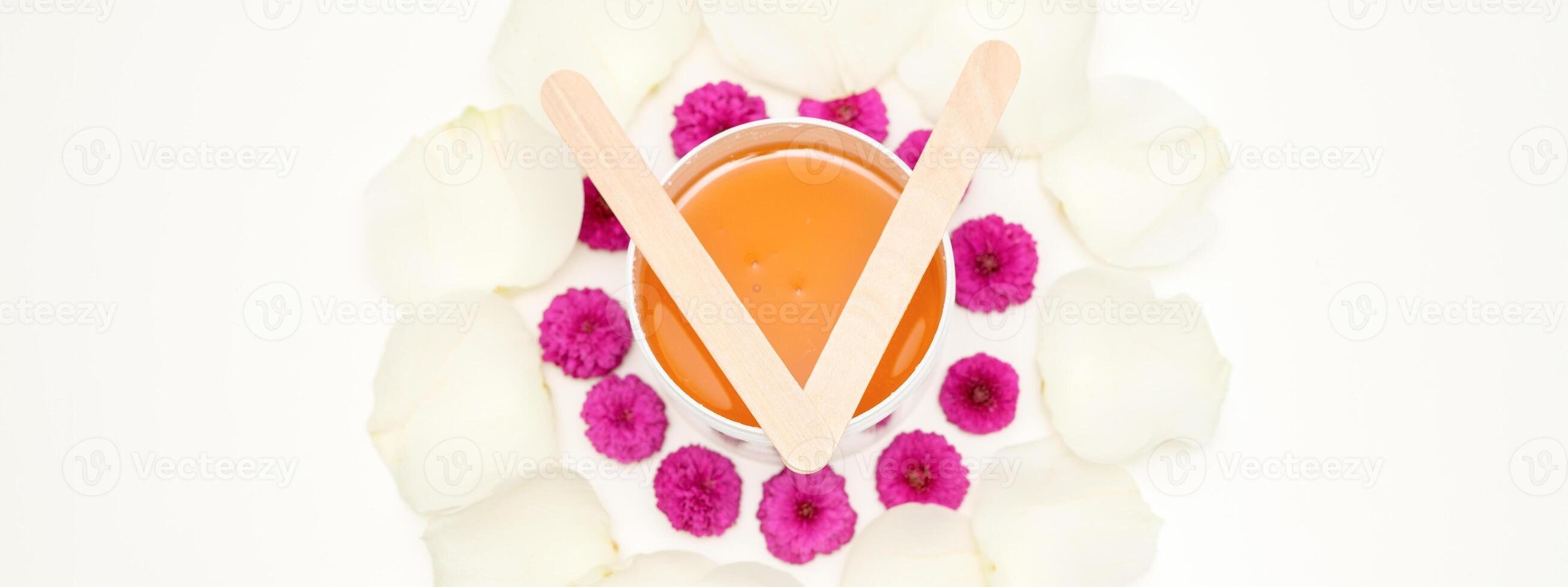 Jar of wax with wooden sticks, pink flowers, and white petals on white background. Concept of depilation. Flat lay top view. photo
