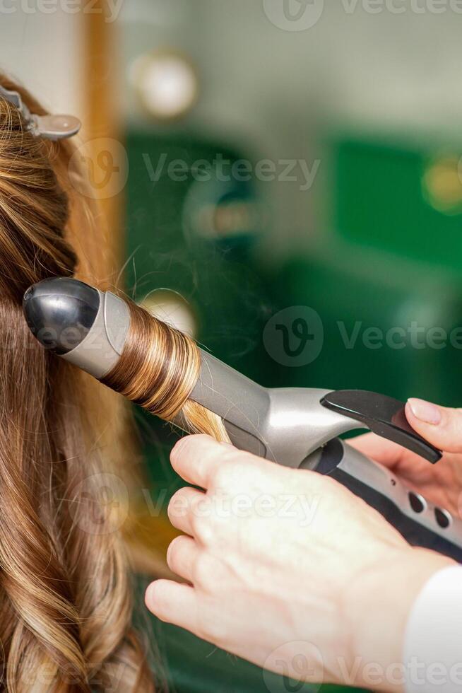 Creating curls with curling irons. Hairdresser makes a hairstyle for a young woman with long red hair in a beauty salon. photo