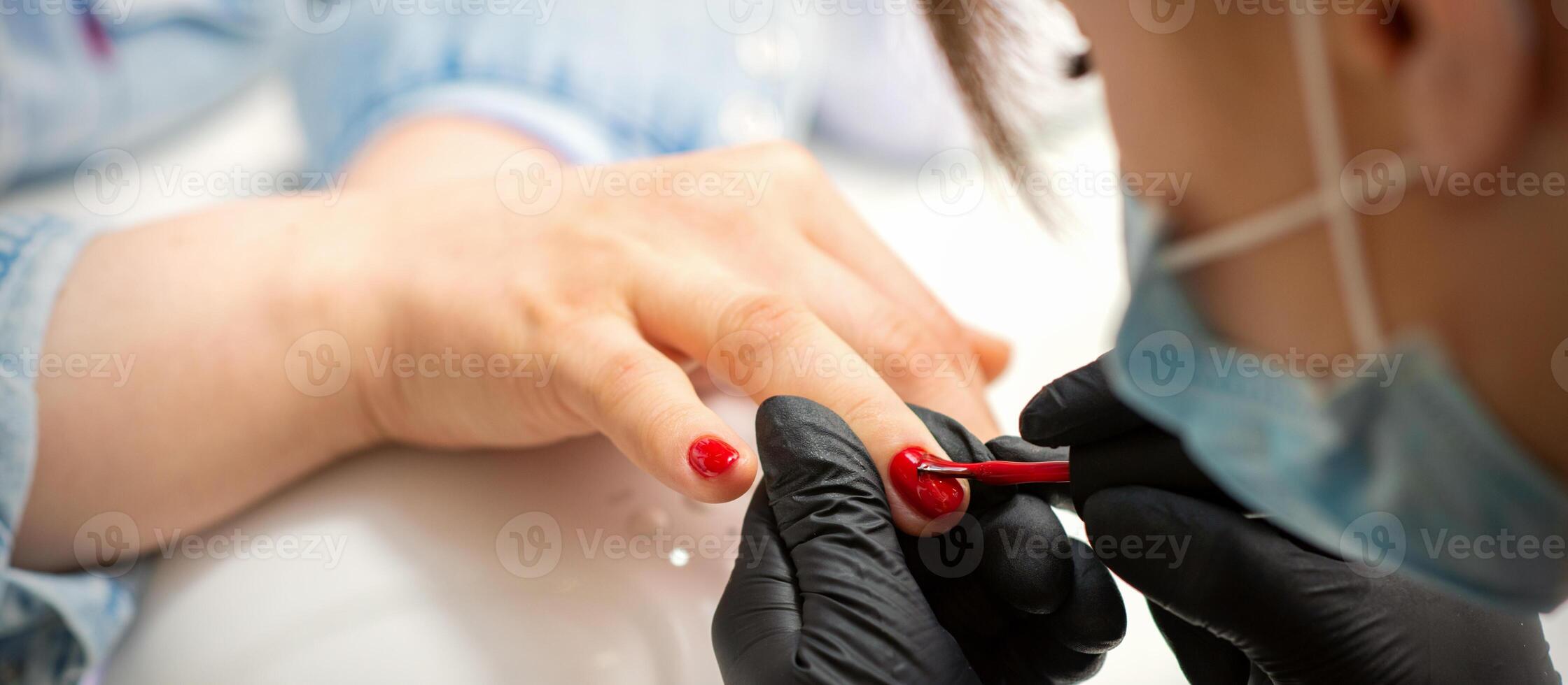Painting nails of a woman. Hands of Manicurist in black gloves applying red nail polish on female Nails in a beauty salon. photo