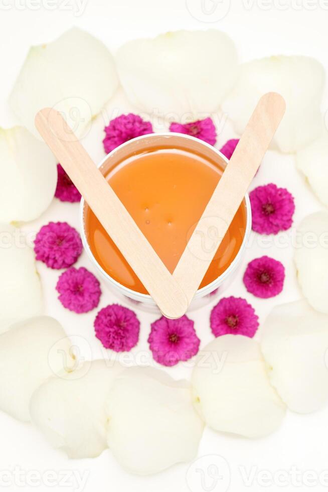 Jar of wax with wooden sticks, pink flowers, and white petals on white background. Concept of depilation. Flat lay top view. photo