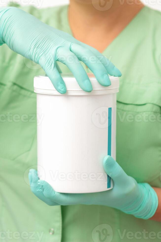 Hands in protective gloves of beautician open a white body cream jar on white background. photo