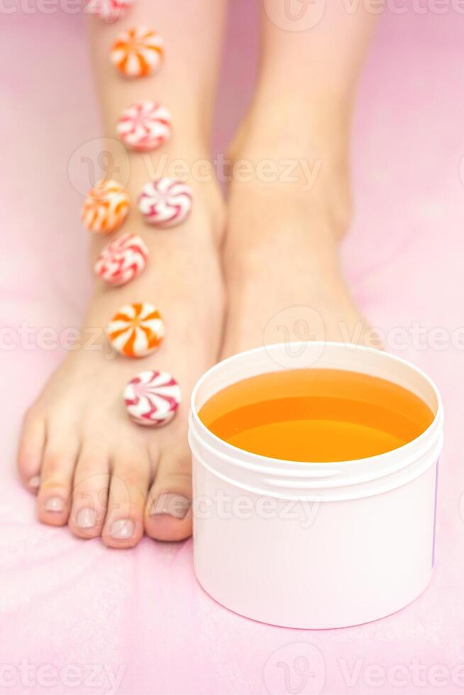 Round candies lying in a row on female feet with white jar with sugar paste on pink background with copy space, depilation concept. photo