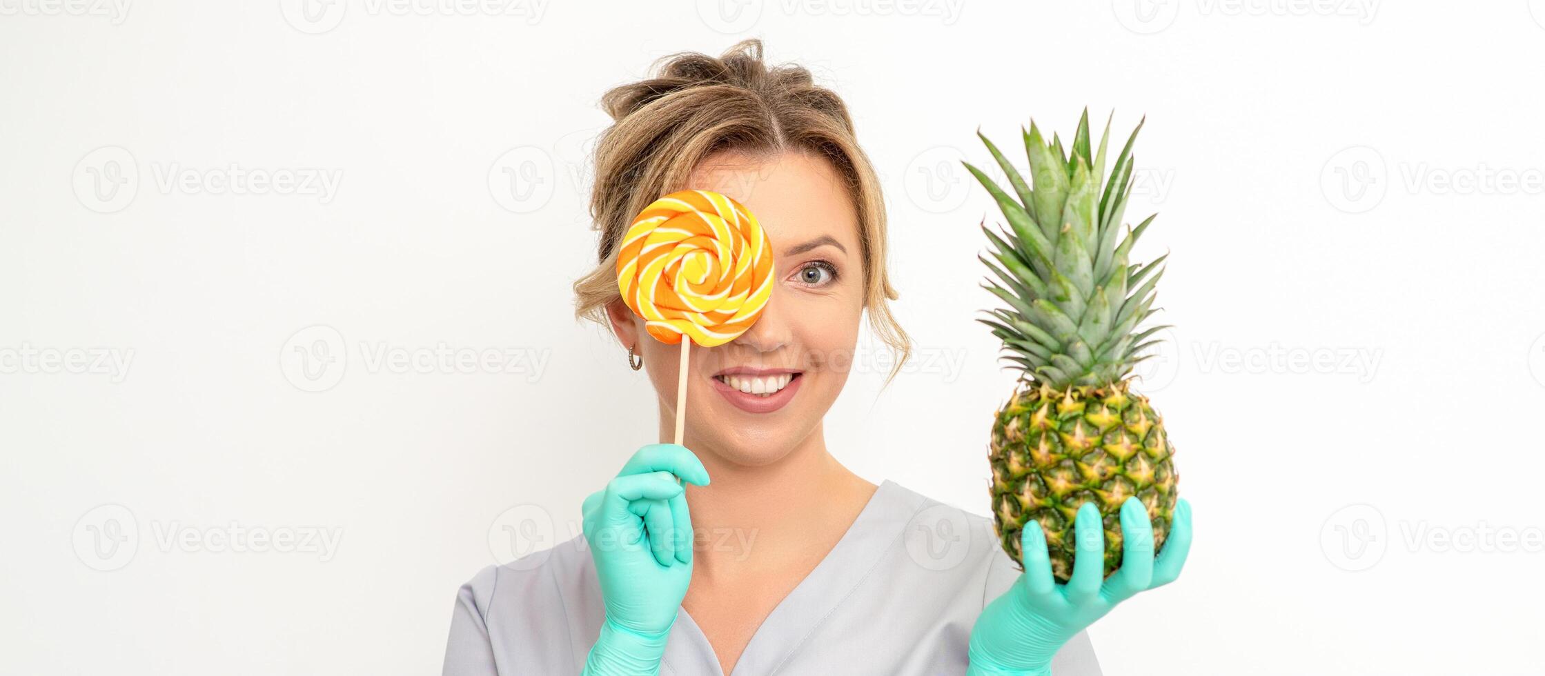 Beautiful smiling beautician holding fresh pineapple and cover her eye with lollipop over white background. Skincare cleansing eco organic. photo