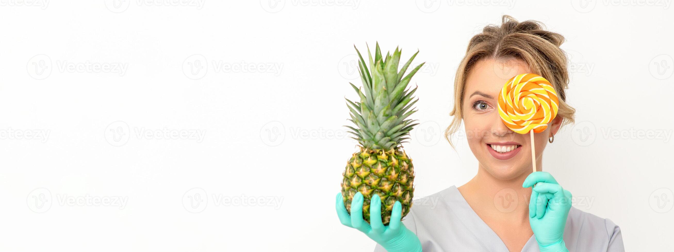Beautiful smiling beautician holding fresh pineapple and cover her eye with lollipop over white background. Skincare cleansing eco organic. photo