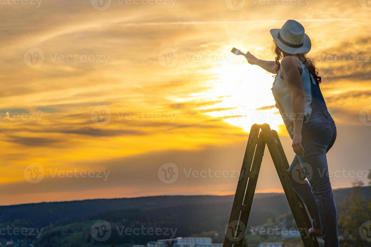 a woman in a work pants stands on a wooden ladder and paints the sunset photo