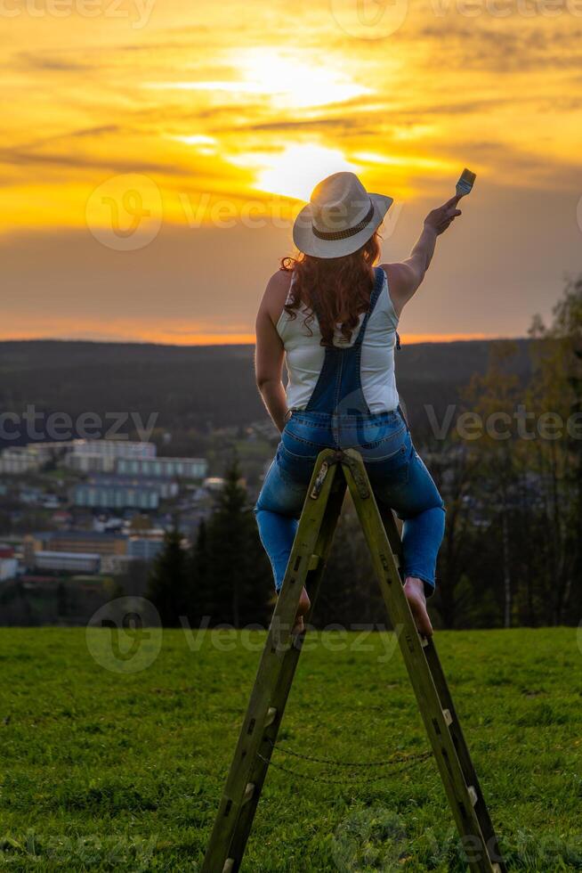 a woman in a work pants stands on a wooden ladder and paints the sunset photo