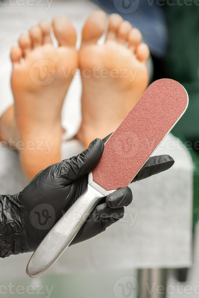 Special grater for scrubbing heels in hands of nail pedicure technician on feet background in beauty salon closeup. photo