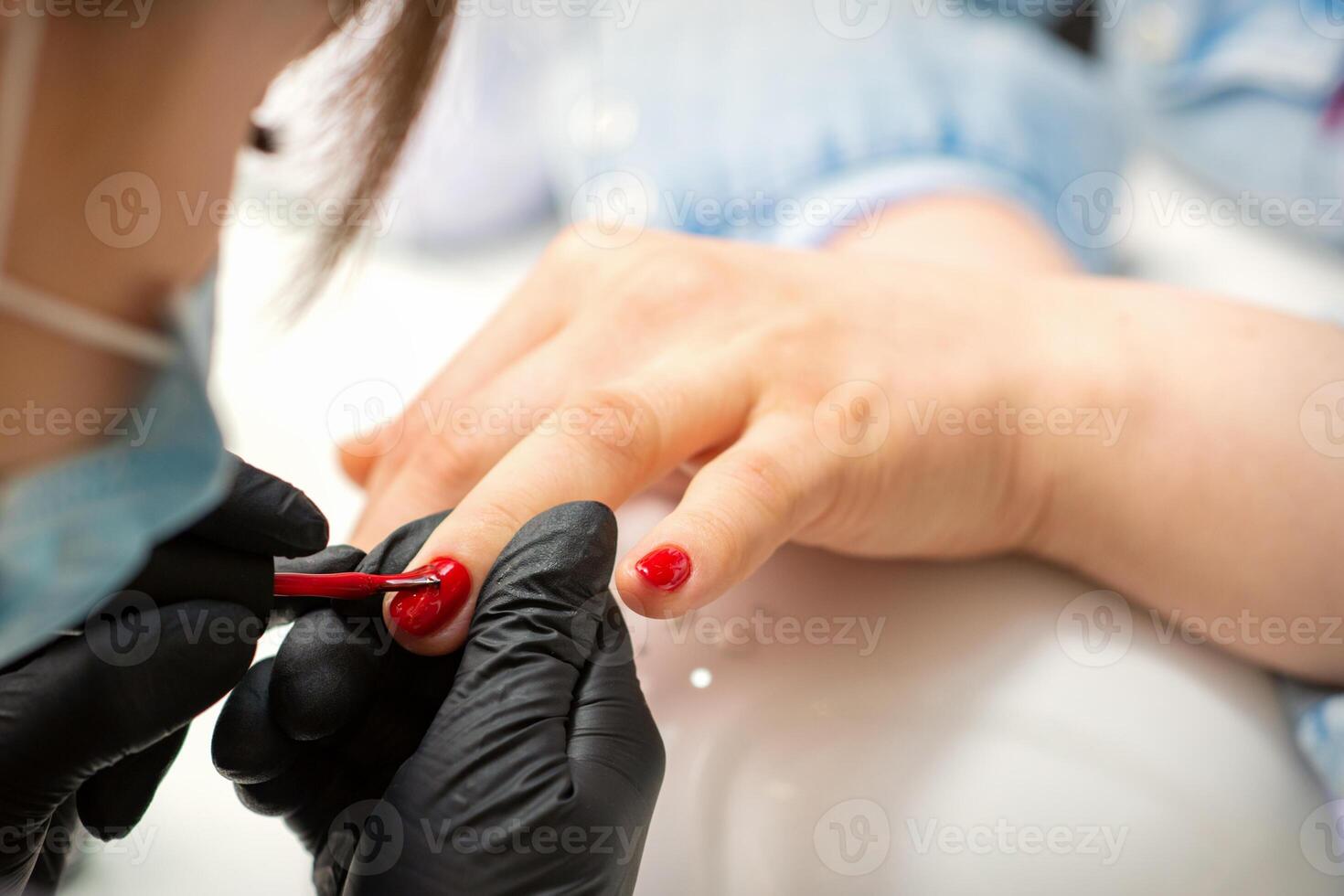 Painting nails of a woman. Hands of Manicurist in black gloves applying red nail polish on female Nails in a beauty salon. photo
