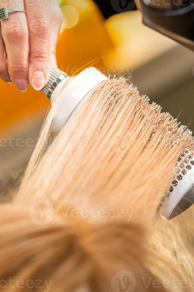 peluquero mano el secado rubio pelo con un secador de pelo y redondo cepillo en un belleza salón. foto