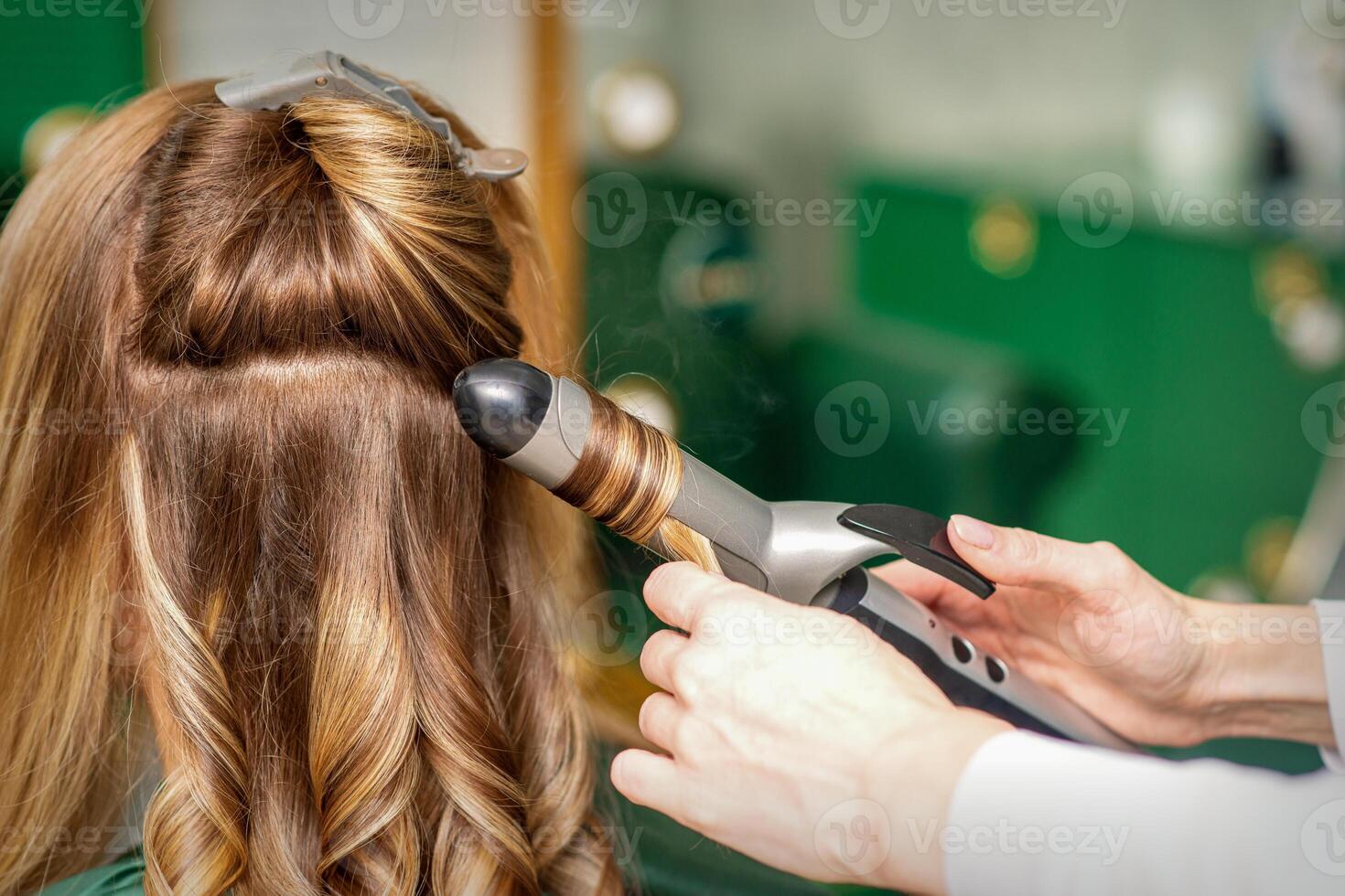 Creating curls with curling irons. Hairdresser makes a hairstyle for a young woman with long red hair in a beauty salon. photo