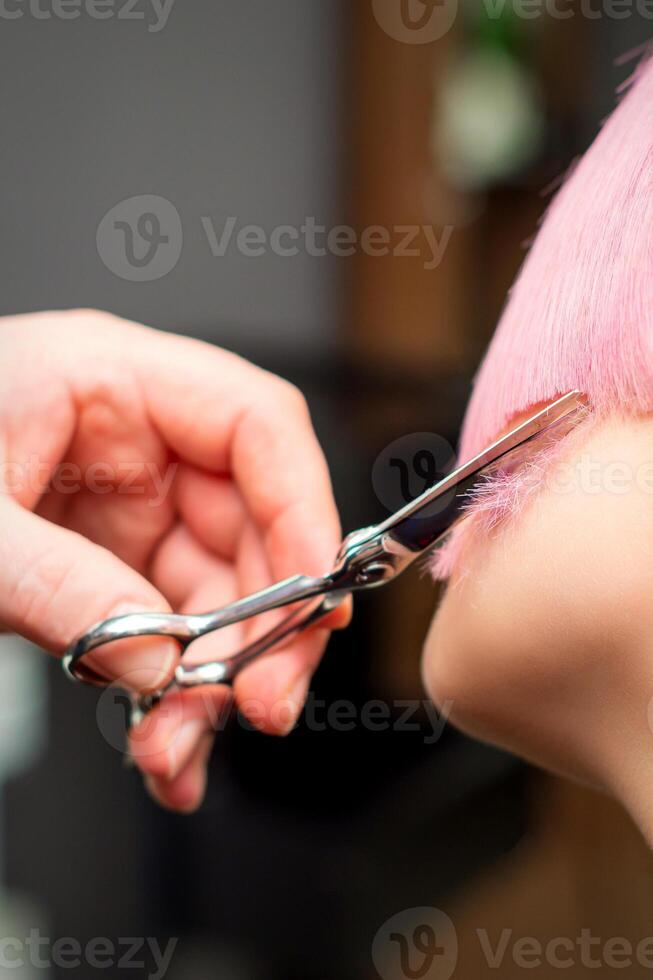 Cutting female hair. Hand of a hairdresser cutting short pink hair of young white woman at the hair salon. photo