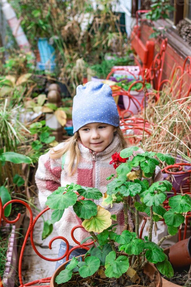 Adorable little girl with blonde hair smiling near a vibrant flower pot in the garden. photo