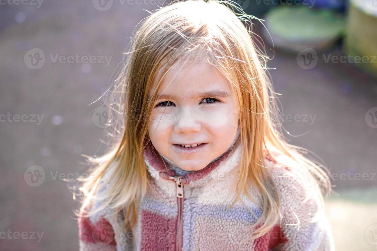 Smiling young girl wearing a stylish checkered jacket, posing for the camera with a blurred outdoor background of trees. She exudes joy and happiness in the natural setting. photo