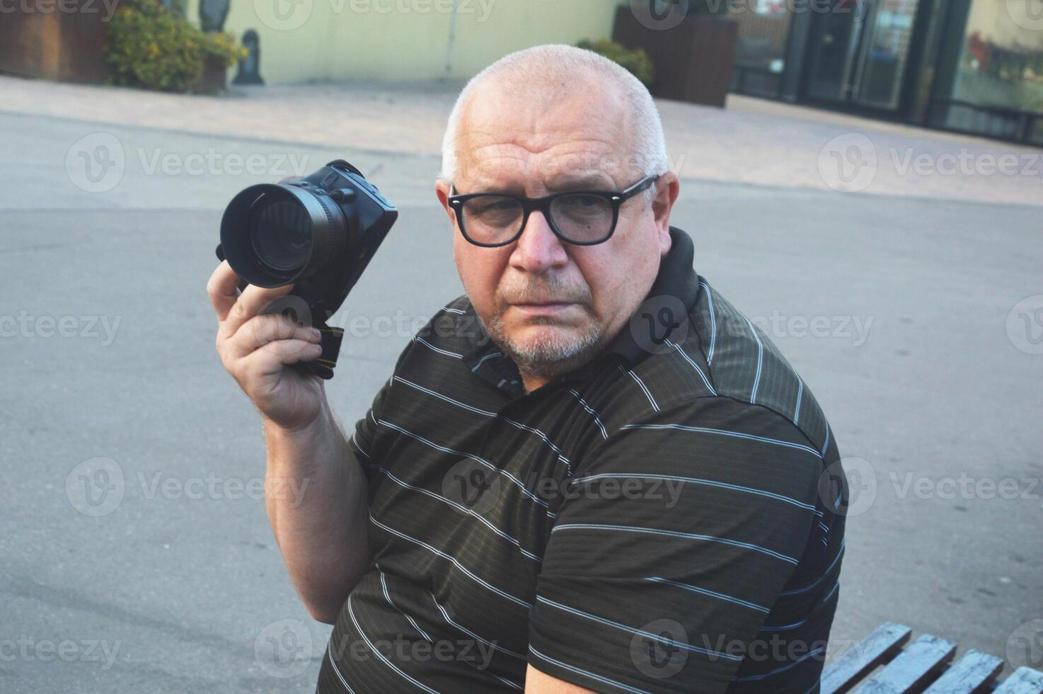 The photographer is an older Caucasian man in glasses sitting on a bench with a modern photo camera.