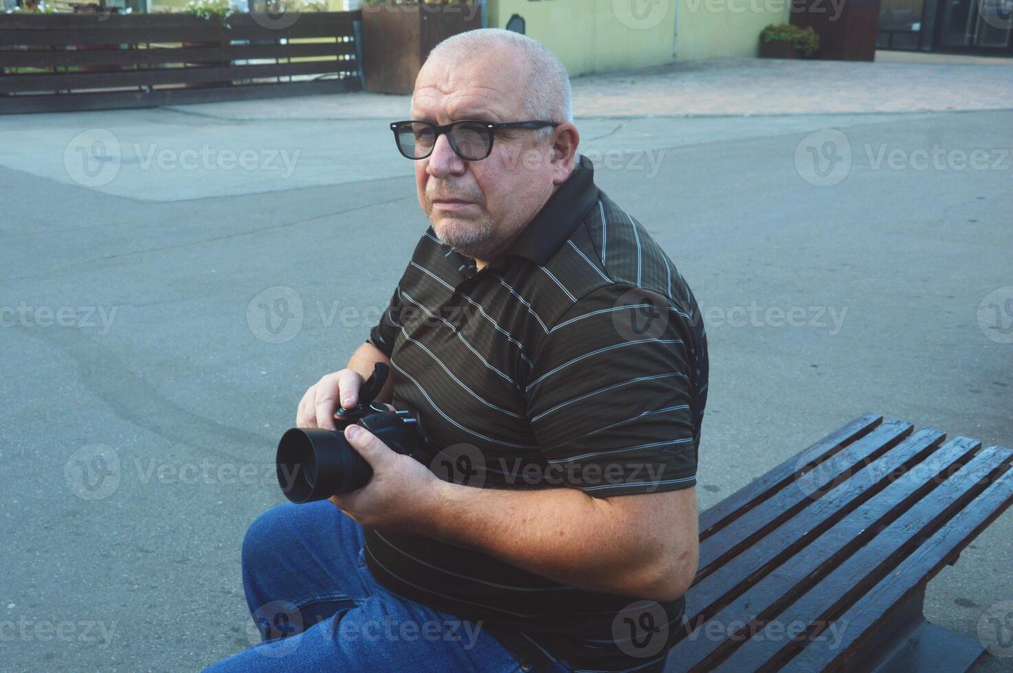 un mayor caucásico masculino fotógrafo en lentes sostiene un cámara en su manos mientras sentado en un banco en un ciudad parque. foto