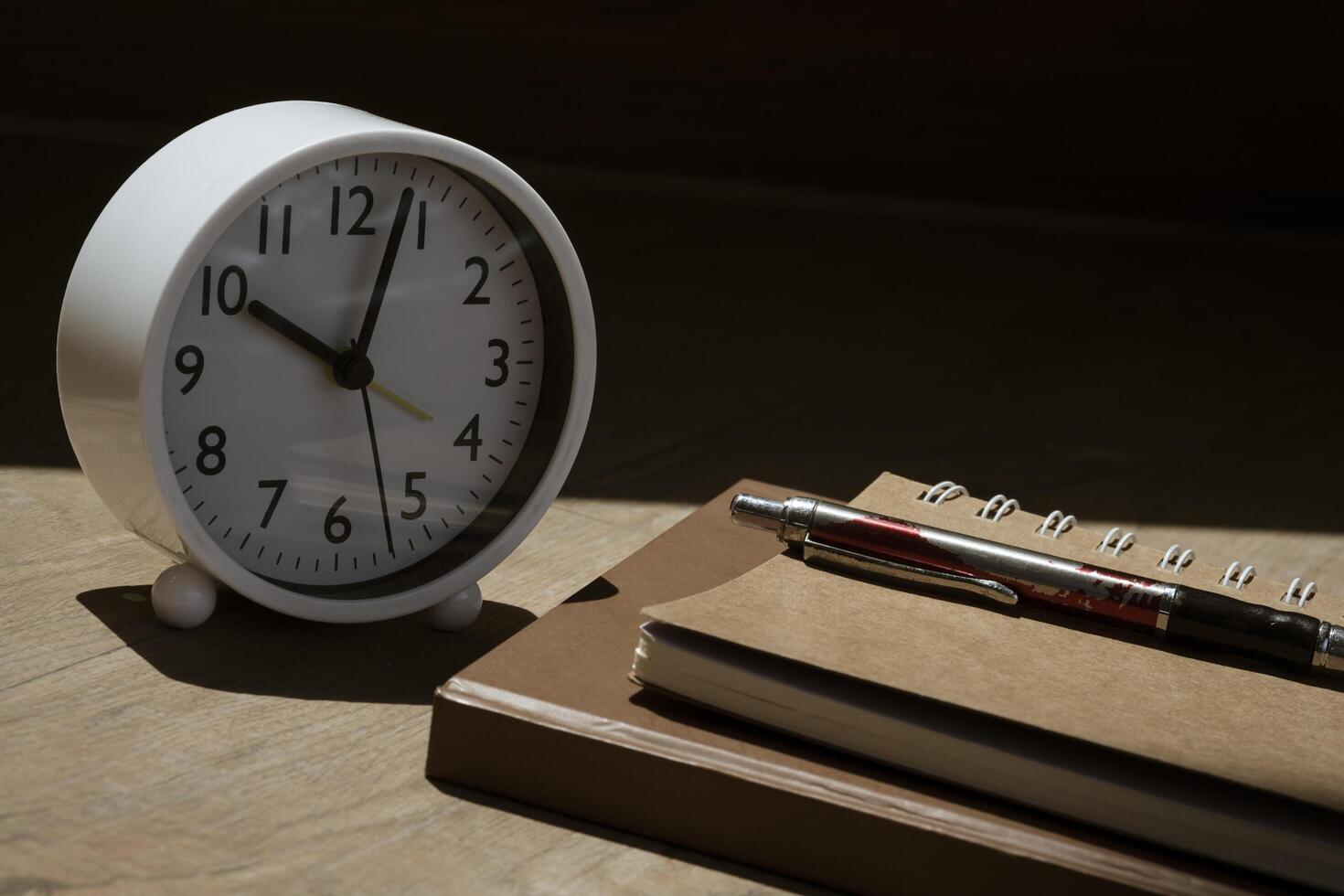 Selective focus at white table clock and ballpoint pen on stack notebooks with sunlight and shadow on wooden table surface photo