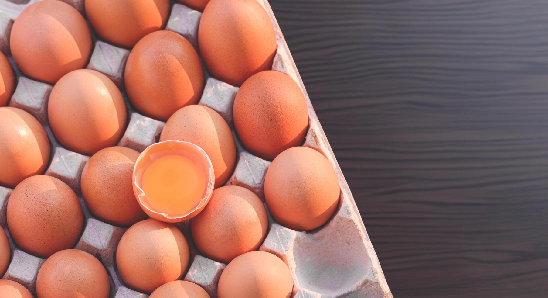 Flat lay of fresh brown Chicken Eggs which one cracked Egg with Yolk in Paper egg Tray on Wooden kitchen Tabletop, top view with copy space photo