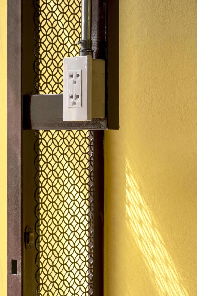 Side view of electrical power socket and conduit pipe line on edge of vintage steel door structure with yellow concrete wall in vertical frame photo