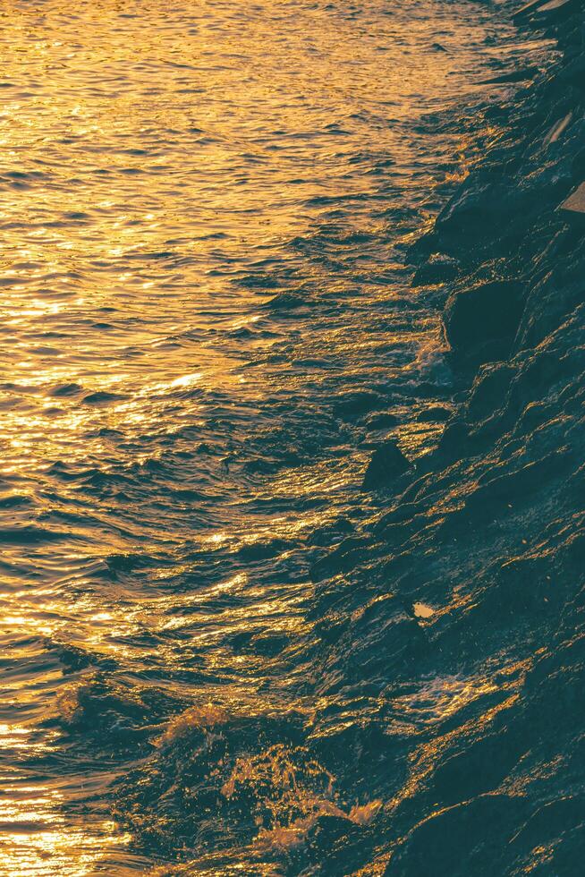 Beautiful golden sunlight reflection on sea surface with rock pile along the beach at sunset time in vertical frame photo