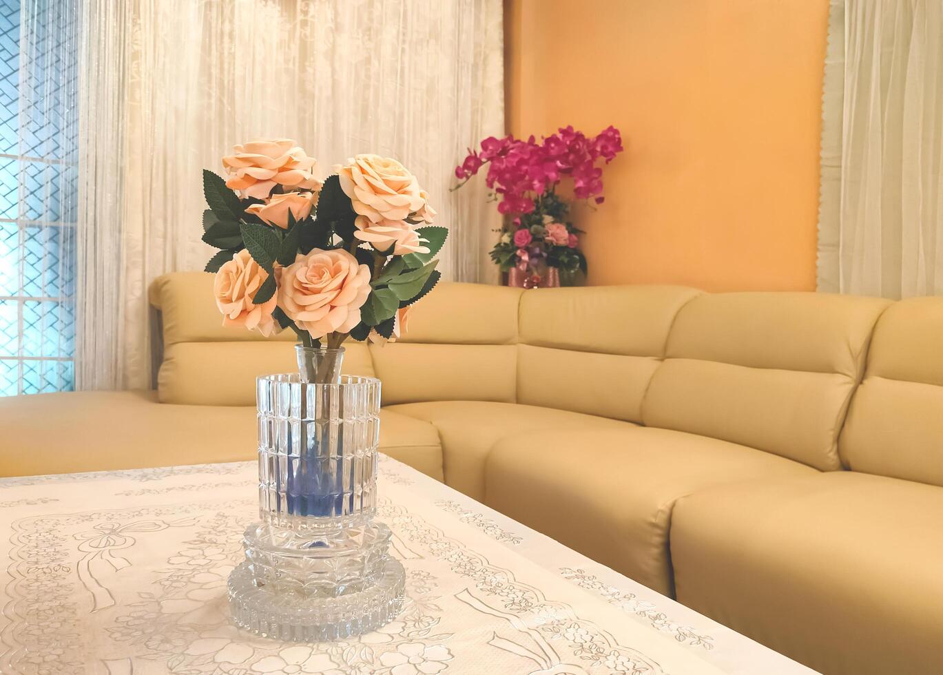 Selective focus at beautiful artificial roses bouquet in crystal vase on a table covered with lace tablecloth with beige sofa in modern living room photo
