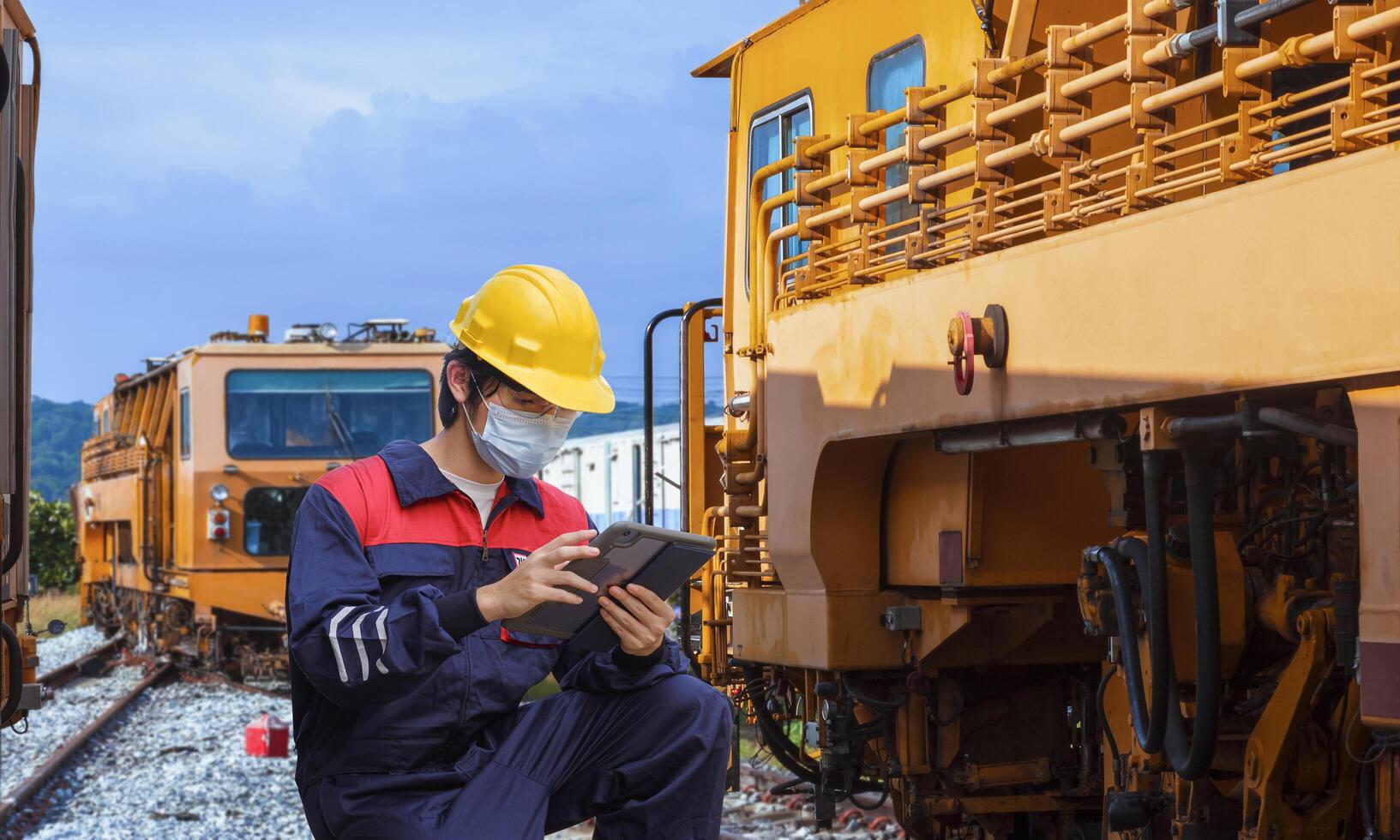 Young Asian engineer in safety workwear with protective mask using digital tablet to record information of the old trains during maintenance at locomotive maintenance station in countryside photo