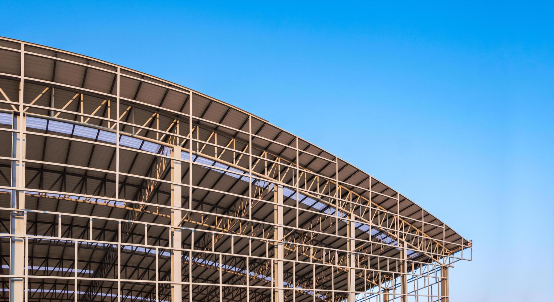 Part of large metal industrial factory building structure with corrugated steel curve roof and skylights in construction site against blue sky background, low angle view with copy space photo
