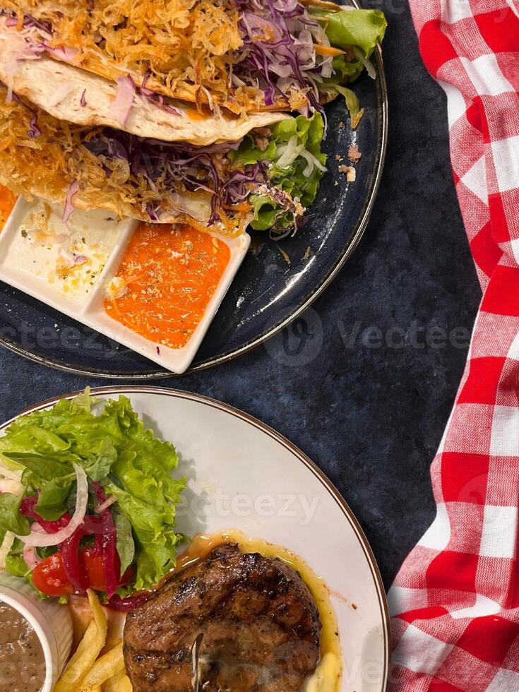 Juicy steak medium rare beef with potato and green salad and veggie tacos on plate over black background. flat lay top view photo