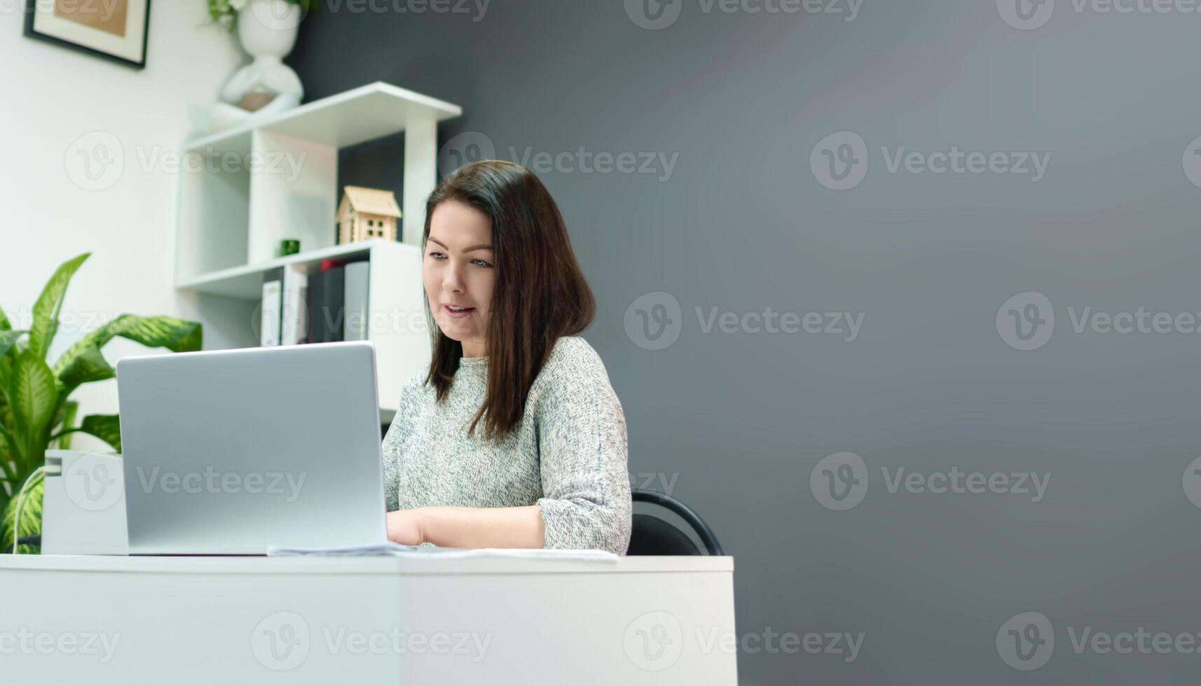 profesional mujer conduce negocio correspondencia mientras sentado a un escritorio con un computadora en un elegante oficina con un gris muro, Copiar espacio foto