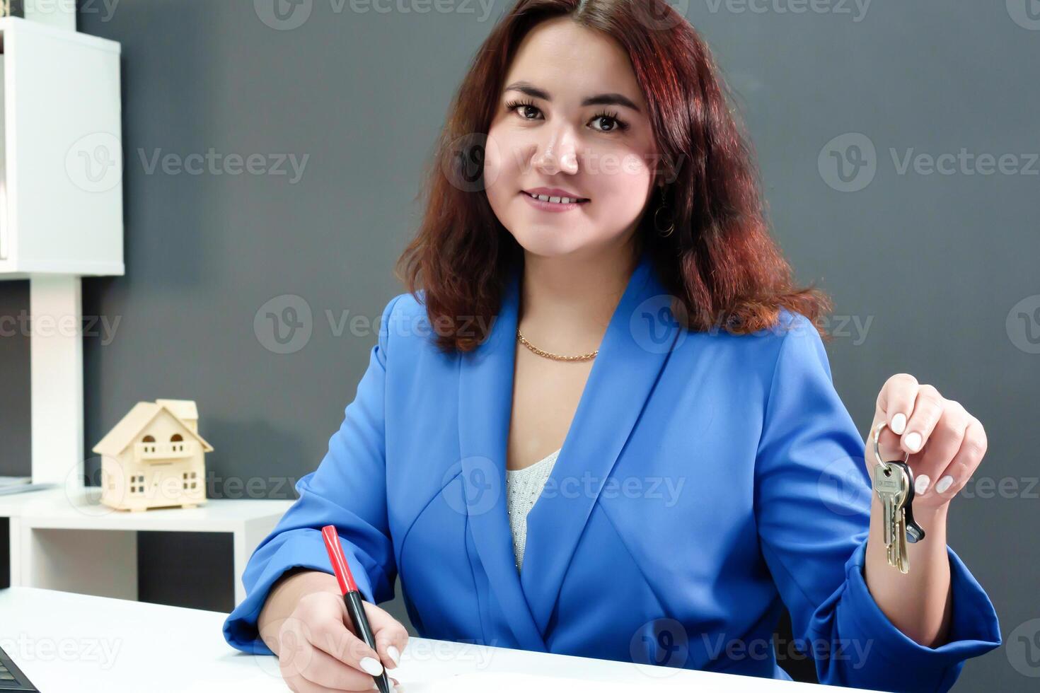 un encantador morena con Rizado pelo y un sonrisa, vestido en un profesional azul traje, sostiene su llaves en su manos a su escritorio, demostrando su papel como un corredor de bienes raíces foto