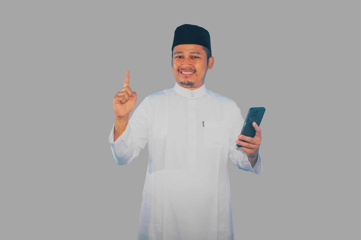 Moslem Asian man smiling happy while holding mobile phone and pointing to the top side photo