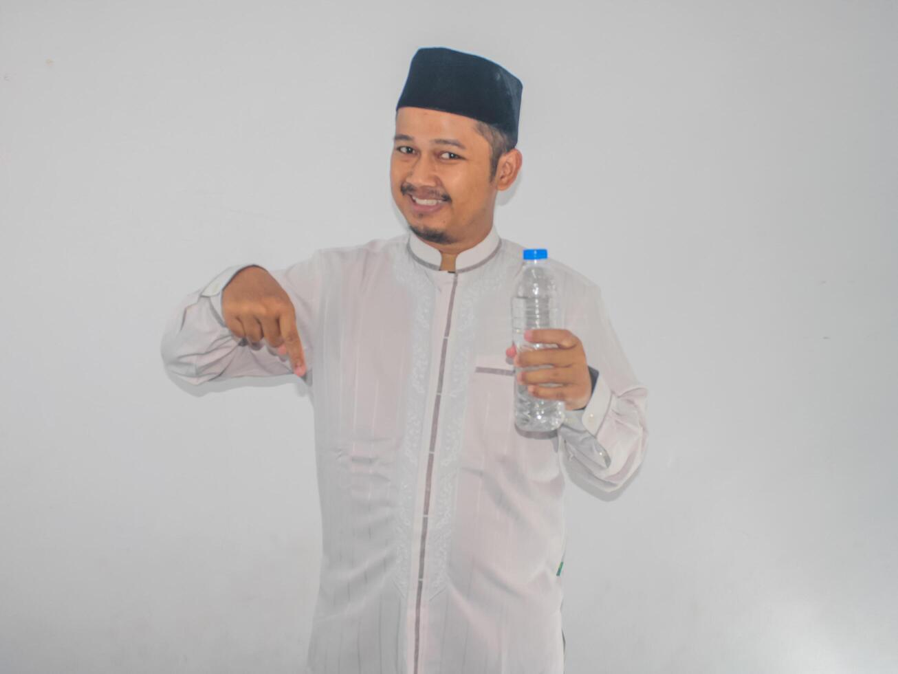 Moslem Asian man smiling and pointing finger down while holding a bottle of drinking water photo