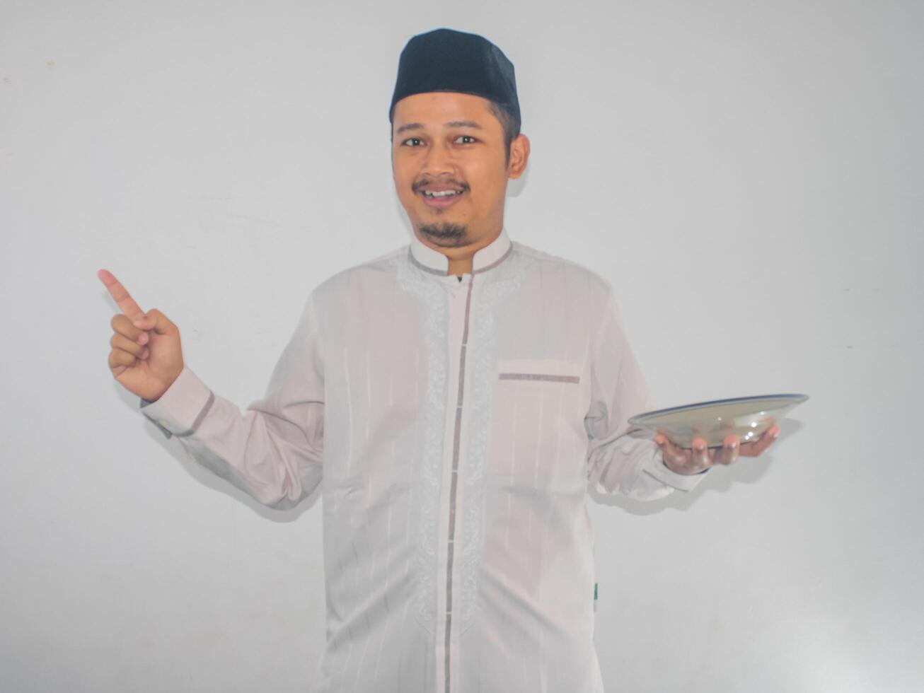 Moslem Asian man smiling and pointing to the right side while holding empty dinner plate photo