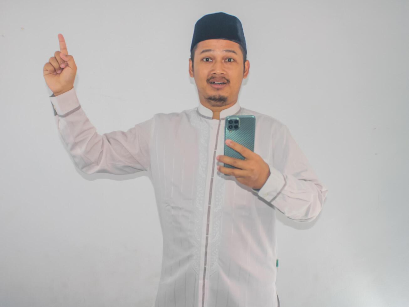 Moslem Asian man shocked while holding mobile phone and pointing to the up side photo