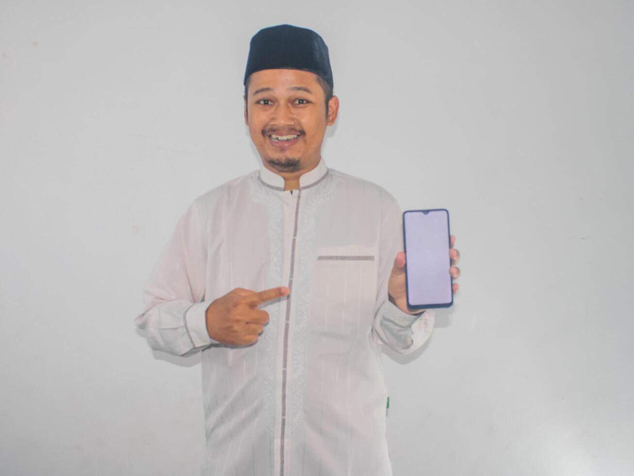 Moslem Asian man smiling and pointing to blank mobile phone screen that he hold photo